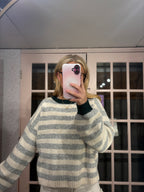 A person taking a mirror selfie wearing a gray and cream striped knit sweater with dark green ribbed collar and cuffs, holding a pink phone in front of their face.
