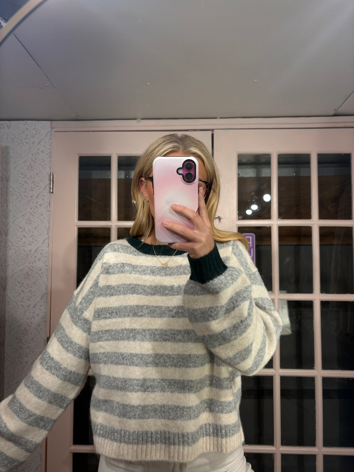 A person taking a mirror selfie wearing a gray and cream striped knit sweater with dark green ribbed collar and cuffs, holding a pink phone in front of their face.