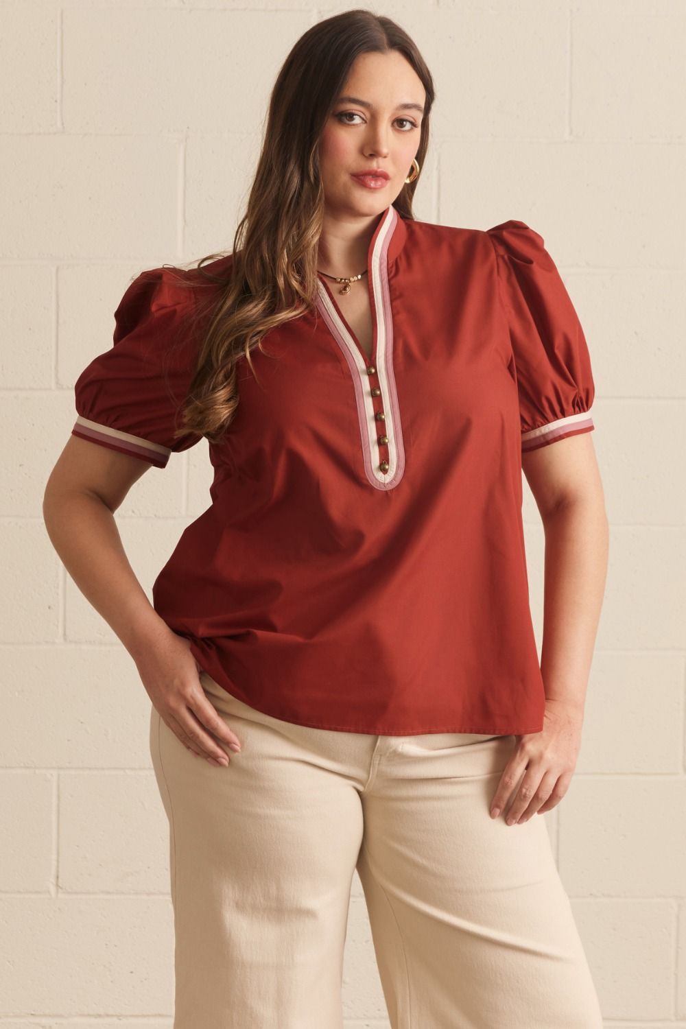 A woman modeling a rust red puff-sleeve blouse with a white and brown trim along the split neckline, paired with cream pants, standing against a light beige brick wall.