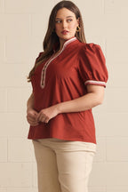 A woman modeling a rust-red puff-sleeve blouse with a white and red trim detail on the front placket, paired with beige pants, standing against a light beige brick wall.