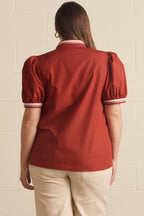 A woman viewed from the back wearing a rust-red short-sleeve blouse with puff sleeves and white striped trim at the collar and cuffs, paired with beige pants, against a light beige brick wall.