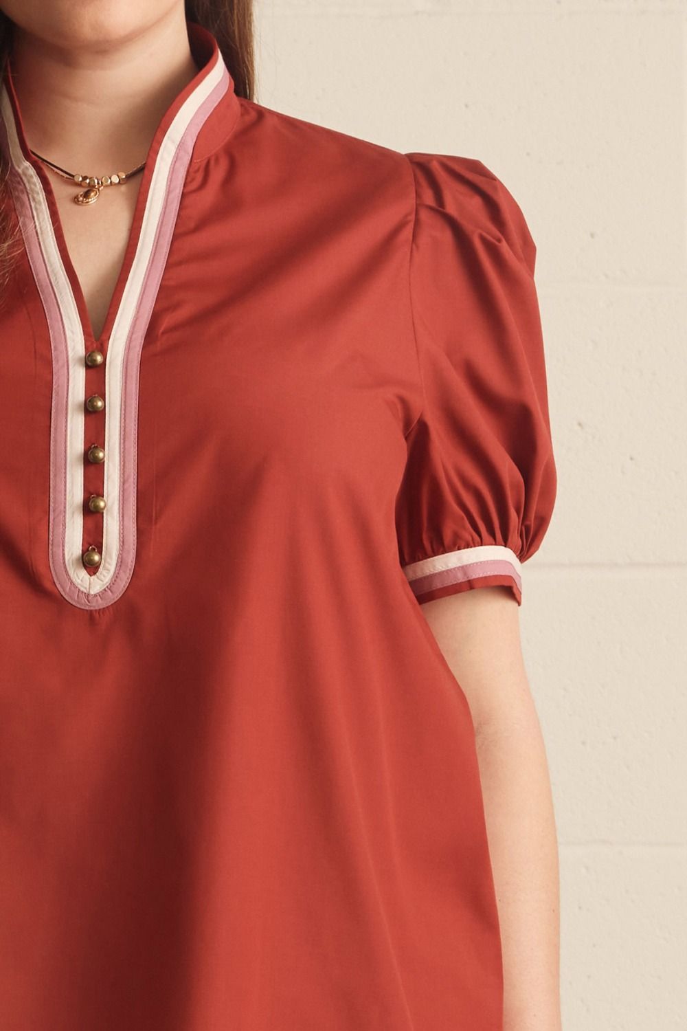 A close-up of a woman wearing a rust-red short-sleeve top with puff sleeves, featuring a V-neckline with white and pink striped trim and decorative gold buttons.