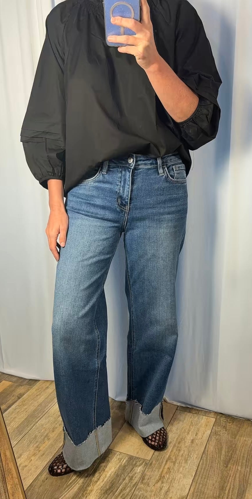 A person taking a mirror selfie wearing a black long-sleeve blouse and wide-leg blue jeans with cuffed hems, standing on a wood floor against a light curtain backdrop.