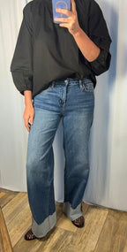 A person taking a mirror selfie wearing a black long-sleeve blouse and wide-leg blue jeans with cuffed hems, standing on a wood floor against a light curtain backdrop.