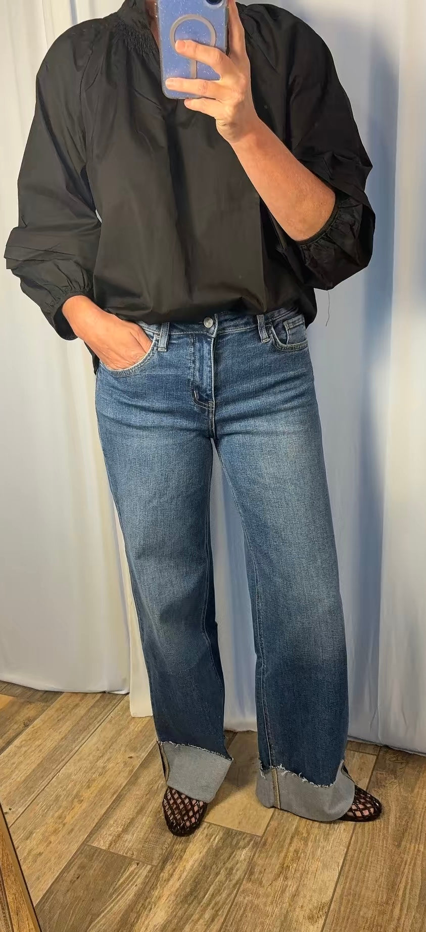 A person taking a mirror selfie wearing a black long-sleeve blouse, blue wide-leg jeans with rolled cuffs, and black-and-white patterned shoes, standing against a white curtain backdrop.