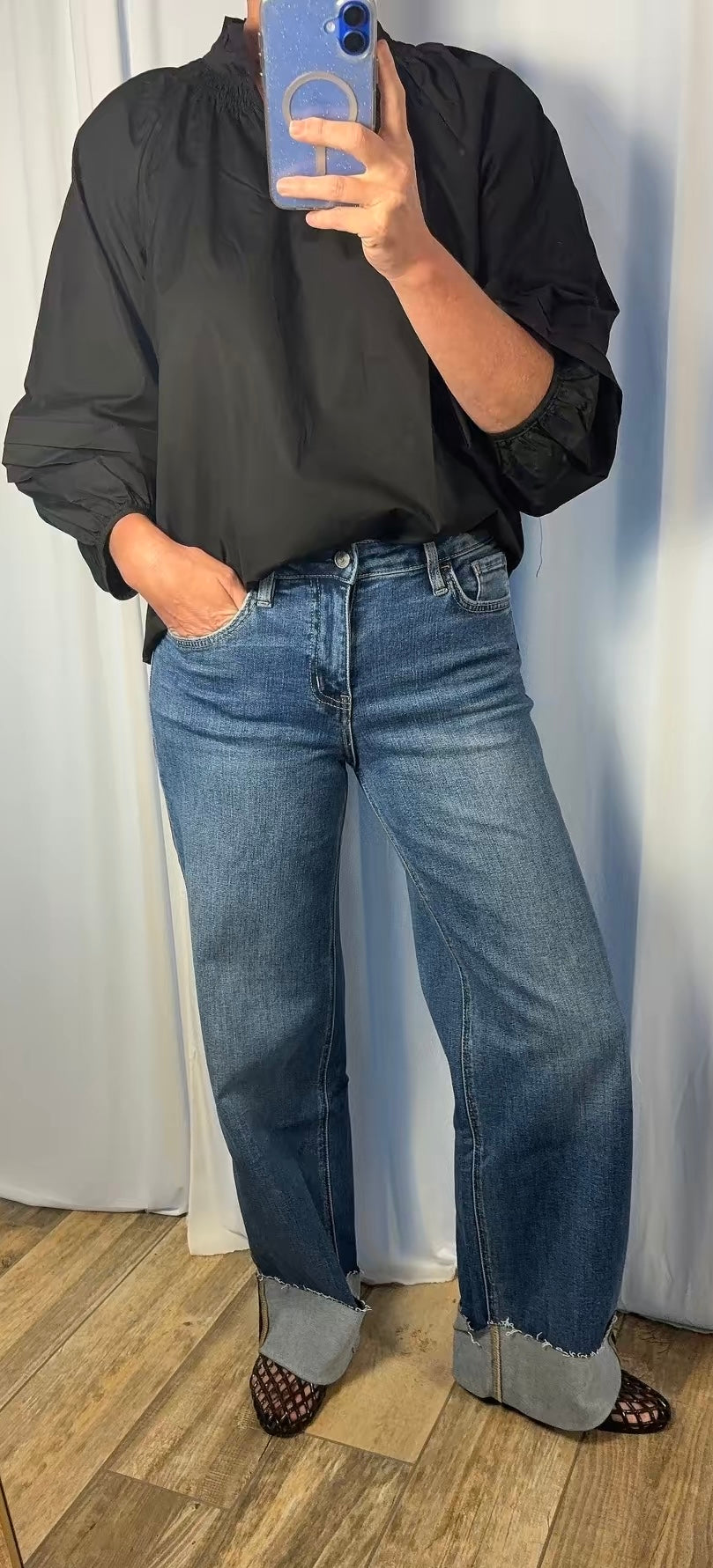A person taking a mirror selfie wearing a black long-sleeve top, blue wide-leg jeans with rolled cuffs, and black-and-white patterned shoes, standing on a wood floor against a white curtain backdrop.